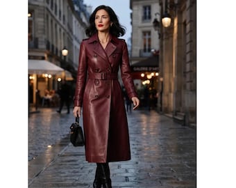 May include: A long, double-breasted trench coat in a deep burgundy leather finish, featuring a notched collar, shoulder epaulets, long sleeves with buckled cuffs, and a matching waist belt with a buckle. A small black handbag is held at the side.