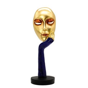 Op de afbeelding: Een goudkleurige gezichtsmasker sculptuur ondersteund door een blauwe arm met goud geschilderde vingers. De sculptuur heeft een zwarte cirkelvormige basis en staat tegen een witte achtergrond. Het masker heeft gesloten ogen en een serene uitdrukking.