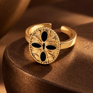 May include: A gold-toned open ring featuring an oval-shaped face with a beaded border. The center of the oval displays an embossed floral-like pattern accented with four smooth, black, petal-shaped inlays arranged in a cross formation.