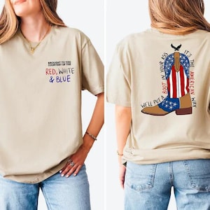 May include: A beige t-shirt featuring a small front graphic that reads BROUGHT TO YOU COURTESY OF THE RED, WHITE & BLUE. The back graphic displays a cowboy boot with an American flag pattern and the text WE'LL PUT A BOOT IN YOUR ASS IT'S THE AMERICAN WAY.