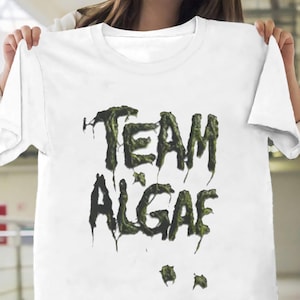 May include: A white short-sleeved t-shirt featuring the text "TEAM ALGAE" printed in a dark green, textured, dripping font that mimics the appearance of slimy algae. The graphic is centered on the chest of the garment, which has a crew neckline.