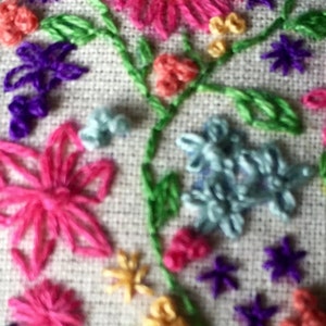 May include: Close-up of a colorful embroidered design featuring flowers, leaves, and stars. The embroidery is done on a white fabric background.