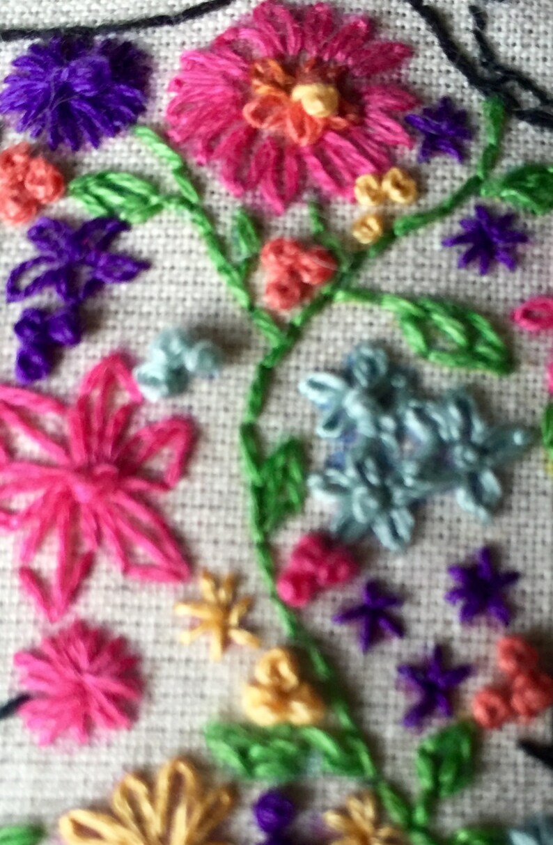 May include: Close-up of a colorful embroidered design featuring flowers, leaves, and stars. The embroidery is done on a white fabric background.