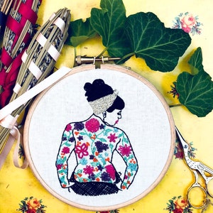 May include: Embroidered hoop art featuring a woman with a floral patterned dress. The woman has dark hair and is wearing a silver headband. The hoop is made of wood and has a white background.
