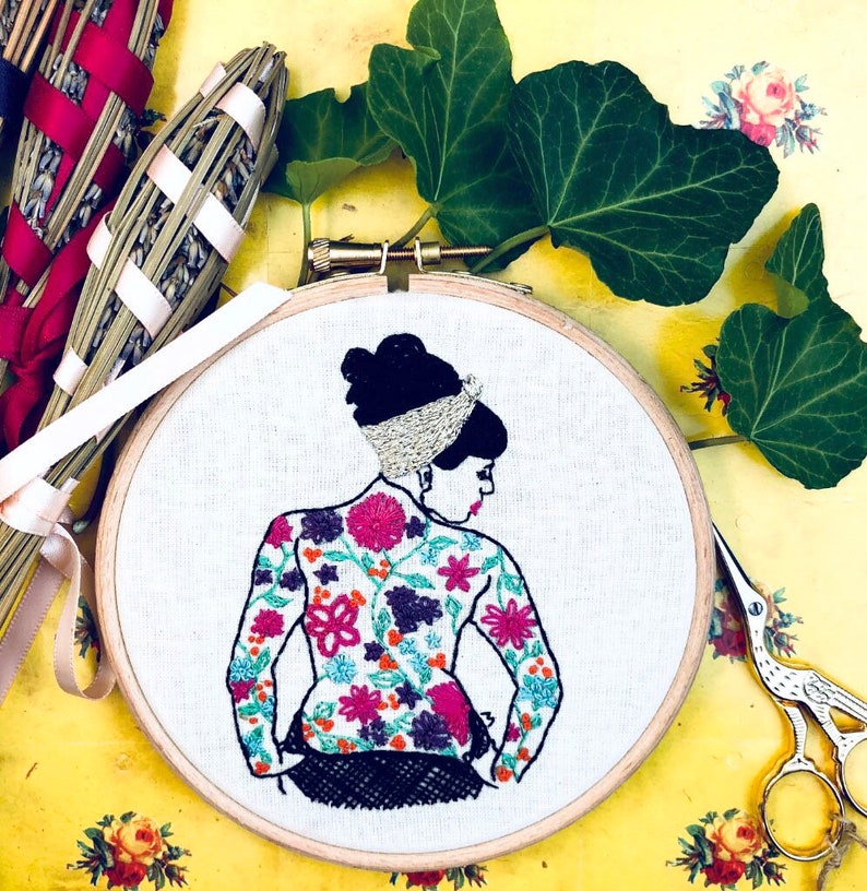 May include: Embroidered hoop art featuring a woman with a floral patterned dress. The woman has dark hair and is wearing a silver headband. The hoop is made of wood and has a white background.