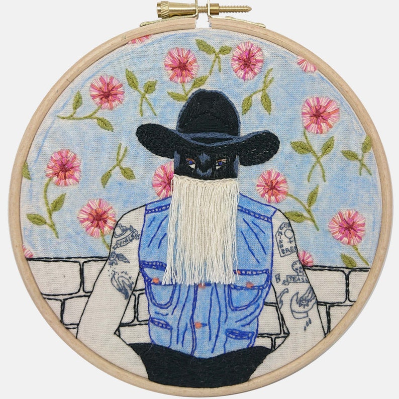 K&ouml;nnte beinhalten: Stickerei eines Menschen mit einem schwarzen Cowboyhut und einer blauen Jeansweste mit einem wei&szlig;en Fransenbart. Der Hintergrund ist ein blauer Himmel mit wei&szlig;en Blumen.