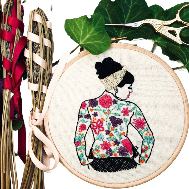 May include: Embroidered hoop art featuring a woman with a floral design on her back. The flowers are in shades of pink, purple, orange, and green. The woman is wearing a black dress and has a gold headband.