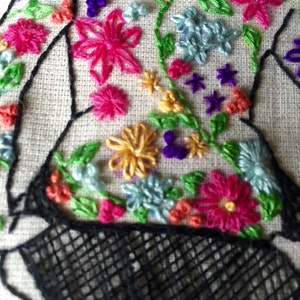 May include: Embroidered floral design on a white fabric background. The design features a variety of flowers in pink, yellow, blue, purple, and green. The flowers are arranged in a random pattern and are surrounded by green leaves. The design is outlined in black thread.