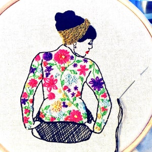 May include: Embroidered hoop art featuring a woman with a floral patterned jacket and a gold headband. The woman is sitting with her back to the viewer and her head is turned to the side. The embroidery is done in a variety of colors, including pink, purple, blue, and green.