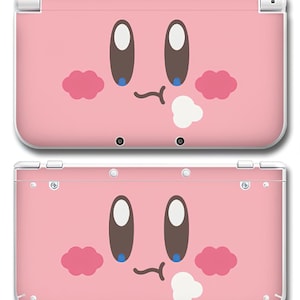 May include: Pink Nintendo 3DS console featuring a cartoon character face design. The face has large brown eyes, pink cheeks, and a small, curved mouth. The design includes white accents and a white puff of air.