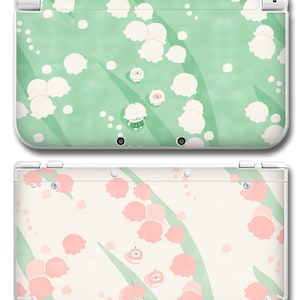 May include: Two Nintendo 3DS consoles, one in light green with white floral designs and the other in cream with pink floral designs. The consoles have a white frame and are decorated with a repeating pattern of flowers and leaves.