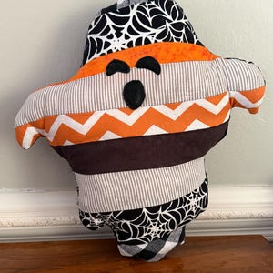 May include: A ghost-shaped stuffed fabric decoration featuring horizontal patchwork panels. The design includes black and white spiderweb patterns, solid orange, grey and white pinstripes, a white and orange chevron stripe, and a solid brown band.
