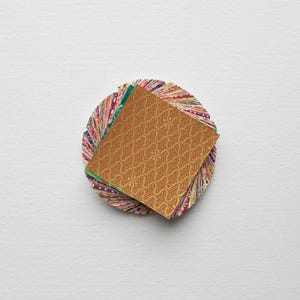 Può includere: Un artigianato circolare in carta multicolore con una pila di carta fantasia sopra. La carta superiore è marrone caldo con un motivo a onde ripetute. La base è composta da molte piccole strisce di carta colorata.