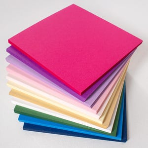 Pode incluir: Uma pilha de folhas de papel quadradas e planas em várias cores, incluindo rosa choque, lavanda, rosa claro, creme, branco, verde floresta, azul brilhante e azul marinho, dispostas em uma pilha escalonada sobre um fundo branco.