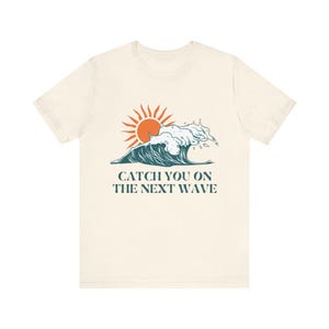 May include: Cream-colored t-shirt featuring a graphic of a stylized ocean wave and sun in orange and teal. The text "CATCH YOU ON THE NEXT WAVE" is printed below the graphic in teal. The shirt has short sleeves and a crew neck.