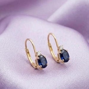 May include: A pair of gold leverback earrings, each featuring a central oval-cut deep blue sapphire gemstone accented by a single small clear round-cut diamond set directly above the sapphire in a polished gold metal setting.