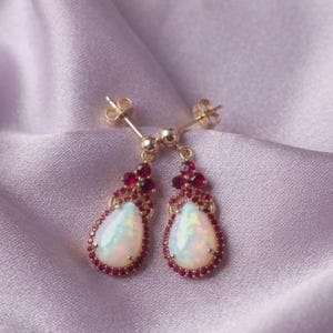 Peut inclure: Une paire de boucles d'oreilles pendantes en or avec des opales en forme de poire entourées de petites pierres précieuses rouge foncé. Les boucles d'oreilles ont une tige et une monture en or délicates, les opales affichant des reflets irisés.