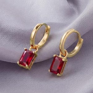 May include: A pair of gold hoop earrings, each featuring a single rectangular-cut, deep red gemstone pendant suspended from the bottom. The metal has a polished finish, and the stones are set in a simple four-prong setting, creating a classic look.