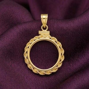Pode incluir: Um pingente dourado com uma moldura circular e uma borda decorativa em forma de corda. O pingente tem uma argola na parte superior para uma corrente e está sobre um fundo de tecido roxo escuro.