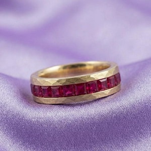 May include: A gold band ring featuring a channel setting of square-cut, vibrant red rubies. The metal surface of the ring displays a textured, hammered finish, contrasting with the smooth, polished edges of the deep red gemstone channel.
