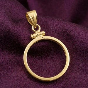 May include: A gold-colored pendant with a circular frame and a decorative bail. The pendant is set against a deep purple fabric background, highlighting its metallic sheen. The bail features a textured design, adding detail to the jewelry.