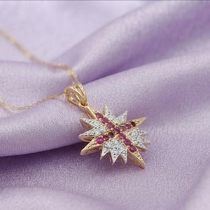May include: A gold pendant necklace featuring a star-shaped design. The pendant is encrusted with small, clear white stones and a central cross pattern of vibrant red stones. The piece hangs from a delicate gold chain, showcasing a detailed metal setting.