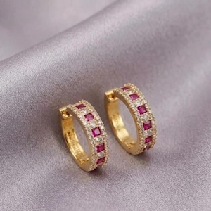May include: A pair of gold hoop earrings featuring a row of square-cut red stones alternating with small clear round stones. The hoops have a hinged closure mechanism and a polished metallic finish, showcasing a symmetrical design.