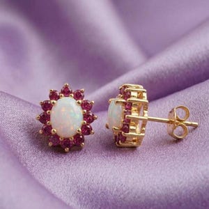 May include: A pair of gold-colored stud earrings. Each earring features a central oval-shaped white gemstone surrounded by small, round, red gemstones. The earrings are set against a purple fabric background.