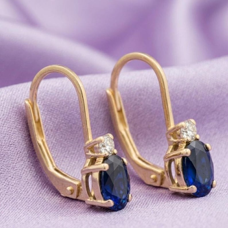 May include: A pair of gold leverback earrings, each featuring an oval-cut deep blue gemstone set in a four-prong mounting, topped with a single small round clear stone. The metal has a polished finish, and the design includes a secure hinged closure.