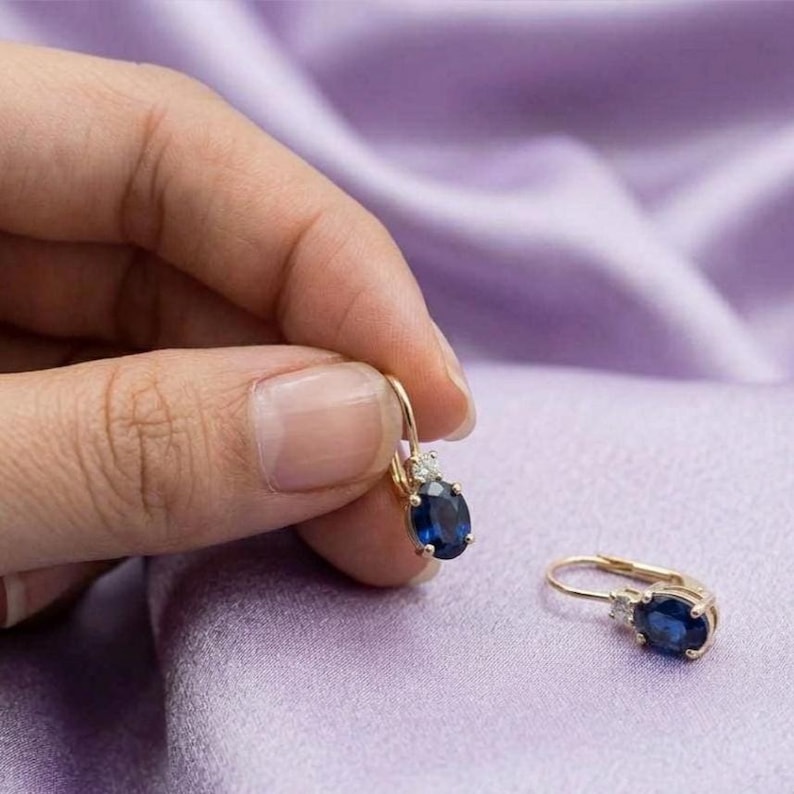 May include: A pair of gold earrings featuring oval-cut blue sapphire gemstones accented by small clear diamonds. Each earring has a secure hinged clasp mechanism, showcasing a deep blue color and a polished metallic finish on the setting.
