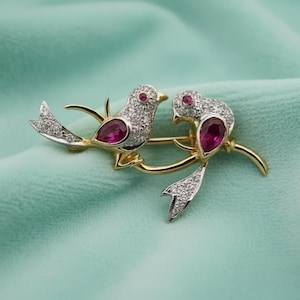 Puede incluir: Broche dorado con dos pájaros posados en una rama. Las aves están adornadas con pequeñas piedras transparentes y detalles de color rubí. El broche se encuentra sobre un fondo suave de color turquesa.