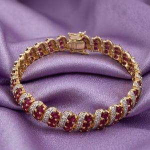 Op de afbeelding: Een goudkleurige armband met afwisselende secties van rode en witte edelstenen. De armband ligt op een paarse satijnen achtergrond. De sluiting is zichtbaar aan de bovenkant van de armband.