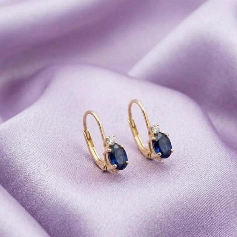 May include: A pair of gold leverback earrings, each featuring a central oval-cut deep blue sapphire gemstone accented by a single small clear round-cut diamond set directly above the sapphire in a polished gold metal setting.