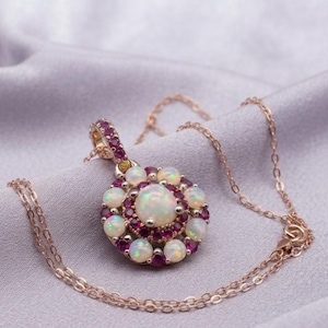 May include: A rose gold pendant necklace featuring a central round opal surrounded by a halo of small round rubies and an outer ring of eight smaller round opals. The pendant is attached to a delicate rose gold chain with a matching metal finish.