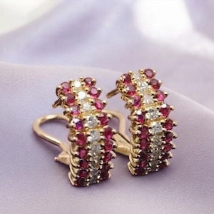 May include: A pair of gold hoop earrings featuring three vertical rows of alternating round-cut red rubies and clear white diamonds. The earrings have a curved, semi-circular design with a secure hinged lever-back closure mechanism for attachment.
