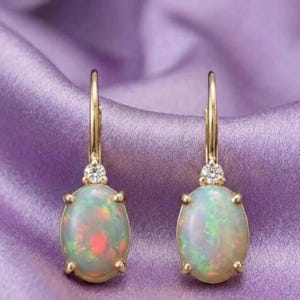 May include: A pair of gold leverback earrings featuring oval-shaped opals with iridescent flashes of red, green, and blue. Each earring is accented with a single small, clear round-cut gemstone set directly above the opal in a prong setting.