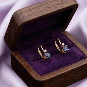 May include: A pair of gold lever-back earrings featuring oval-cut blue sapphire stones and small clear accent diamonds, presented inside a dark brown wooden jewelry box with a deep purple velvet interior lining and a hinged lid design.