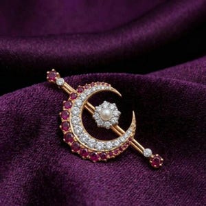 Op de afbeelding: Een goudkleurige halve maan broche met een centrale parel en omringende edelstenen. De maan is afgezet met rode en heldere stenen, en de broche heeft een staafontwerp. De sieraden liggen op een dieppaarse stof.
