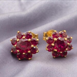 Peut inclure: Une paire de boucles d'oreilles à clous en or avec une pierre précieuse rouge ronde centrale entourée d'un groupe de pierres rouges plus petites en forme de fleur. La monture en or poli maintient chaque pierre rouge individuellement.