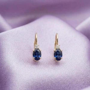 May include: A pair of gold hoop earrings featuring oval-cut deep blue sapphire gemstones suspended from a small gold setting accented with a single clear round-cut diamond at the top of each earring, showcasing a classic drop design.