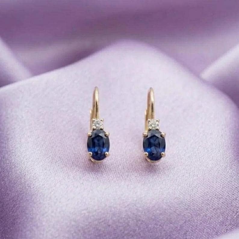 May include: A pair of gold hoop earrings featuring oval-cut deep blue sapphire gemstones suspended from a small gold setting accented with a single clear round-cut diamond at the top of each earring, showcasing a classic drop design.