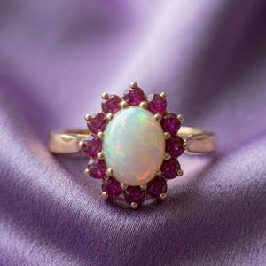 May include: A gold ring featuring a central oval-shaped opal with an iridescent play of colour, surrounded by a halo of twelve round-cut deep pink rubies. The band is smooth and polished, showcasing a classic cluster design with vibrant gemstones.