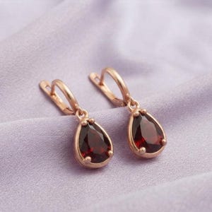 May include: A pair of drop earrings featuring deep red, pear-shaped gemstones set in rose gold metal. Each earring has a hinged lever-back closure, with the faceted stones securely held by small prongs, creating a classic and elegant jewelry piece.