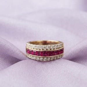 Könnte beinhalten: Ein Goldring mit einer mittleren Reihe aus quadratisch geschliffenen, leuchtend roten Rubinen, flankiert von zwei äußeren Reihen aus kleinen, runden, klaren weißen Diamanten. Der Ring hat eine polierte Metalloberfläche und eine mehrreihige Fassung.
