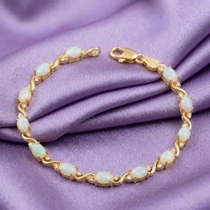 May include: A gold bracelet with oval-shaped opals and small accent diamonds. The bracelet features a delicate, intertwined design. The opals are a creamy white color, and the bracelet is set against a purple background.