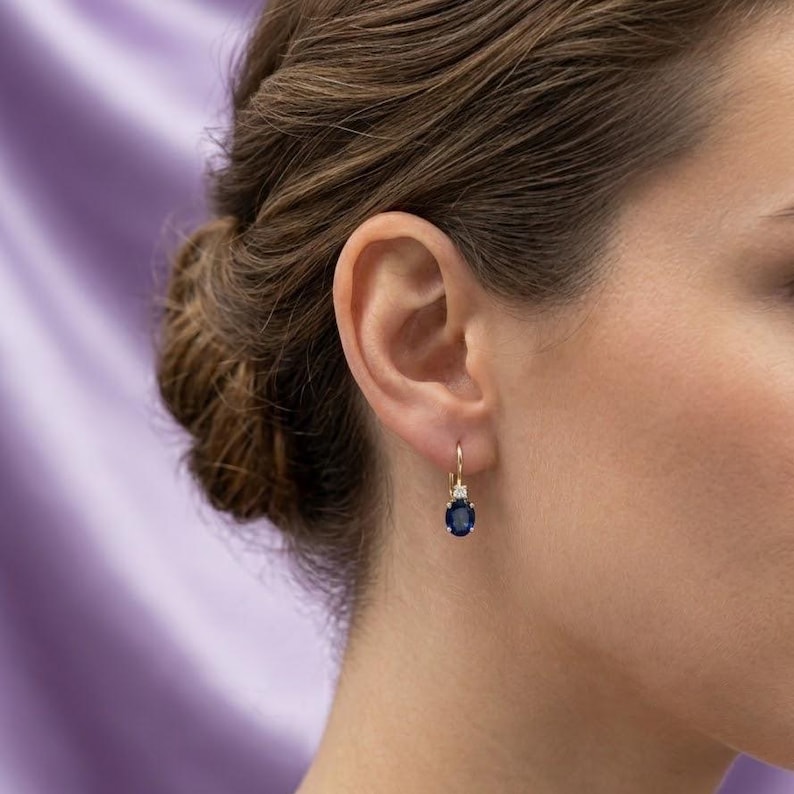 May include: A gold leverback earring featuring a single oval-cut deep blue sapphire gemstone suspended beneath a small, clear, round-cut diamond accent. The earring is worn on the earlobe, showcasing the contrast between the blue stone and gold.