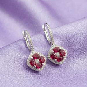 May include: A pair of silver-toned drop earrings featuring a square-shaped pendant. Each pendant is set with four oval-cut deep red stones arranged in a cluster, surrounded by a border of small clear stones and attached to a hinged lever-back closure.