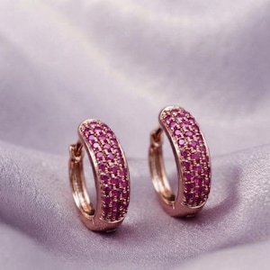 May include: A pair of rose gold hoop earrings featuring three rows of small, round, vibrant pink gemstones embedded along the front surface of each hoop. The earrings have a hinged closure mechanism and a polished metallic finish on the sides.