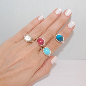 May include: A hand wearing four oval-shaped rings with different colored stones. The rings are silver and the stones are white, red, blue, and turquoise.