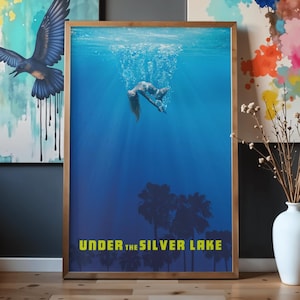 Könnte beinhalten: Ein gerahmter Kunstdruck mit einer Person unter Wasser, umgeben von Blasen, in einem tiefblauen Ozean. Der Druck trägt den gelben Text "UNDER THE SILVER LAKE". Der Rahmen ist hellbraun.